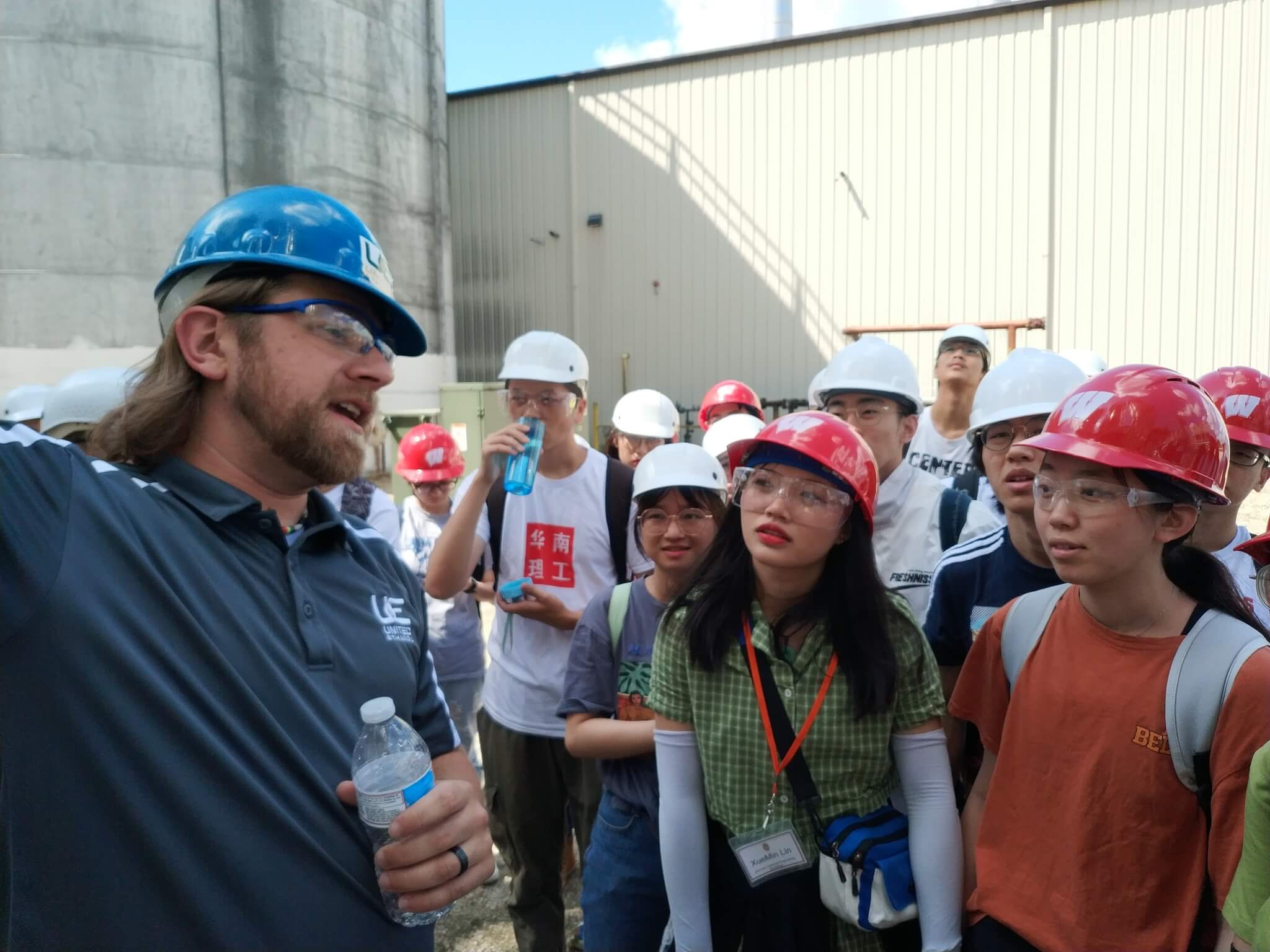 Chinese students touring United Ethanol in Milton; wearing hardhats listening to a speaker standing outside near industrial buildings