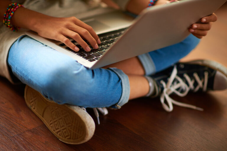 Person typing on a laptop, wearing jeans and sneakers, sitting cross-legged on a wooden floor.