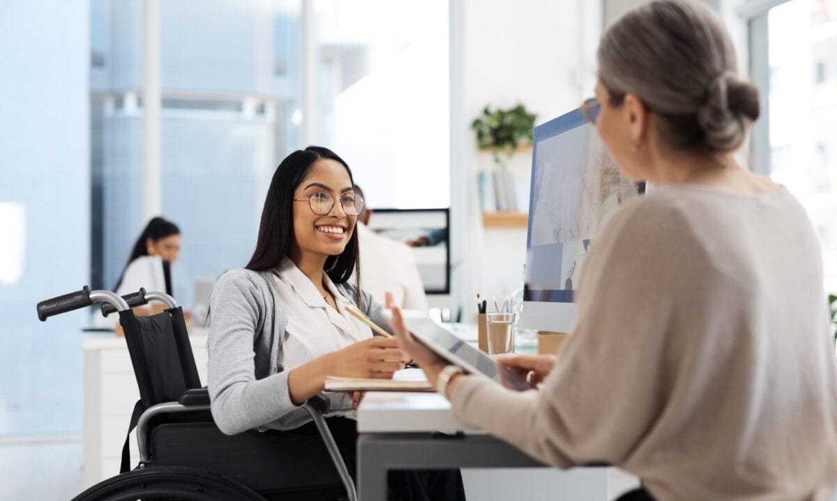 women at desk smiling, sitting in a wheelchair, looking at another woman across from her