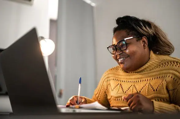 Smiling woman takes notes as she watches laptop screen at home