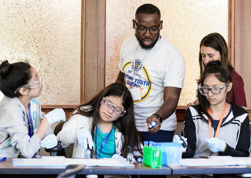 Three middle school students at UW–Madison's Latino Youth Summit sit at a table wearing goggles while conducting a science experiment. Their instructor stands behind them, offering guidance.