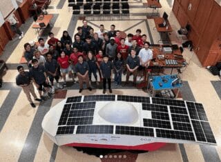 A group of UW–Madison students, photographed from above, stand behind a red and silver solar car. They look up at the camera, smiling.