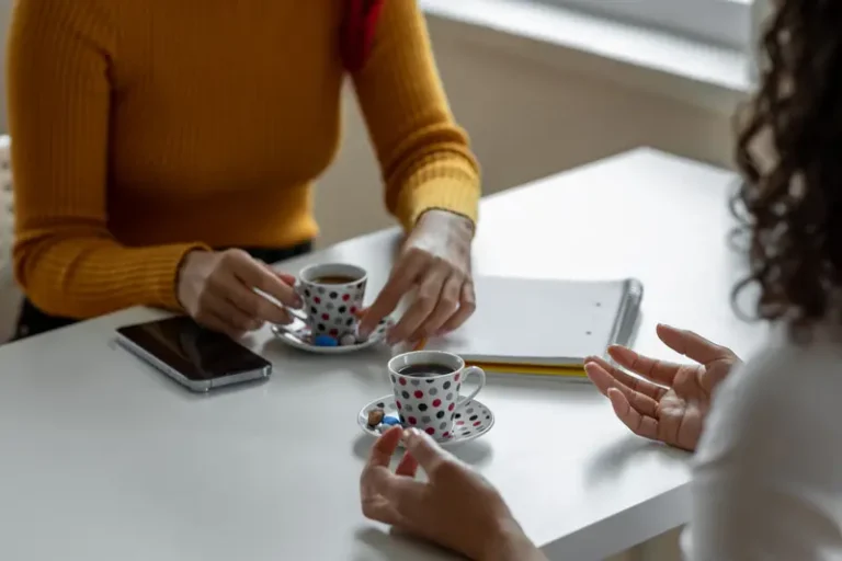 Two people sit facing towards each other for a discussion with notebooks and small coffee cups at a table