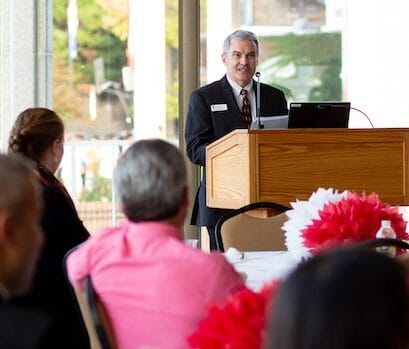 Martin Rouse speaks from a lectern. Audience members are listening while seated at tables in the foreground