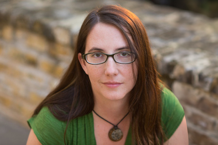 Megan Reilly, wearing a green t-shirt, a glasses and pendant necklace, has long dark reddish brown hair.