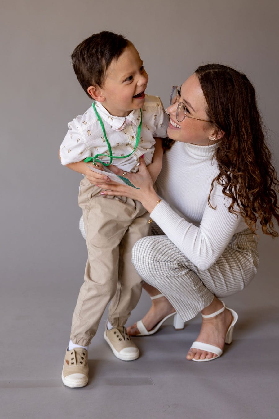 Adult student squatting beside her toddler son tickles his stomach as he laughs, smiling into the camera