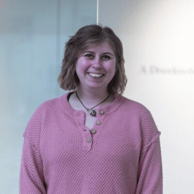 Kagen Dunn smiles at the camera. She has short dark blonde hair and is wearing a pink long-sleeve top and a pendant necklace.