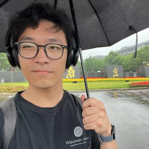 Harry Lou stands in front of a gray stone fence with Chinese characters etched in gold on larger sections. Harry has dark hair and glasses and his holding a black umbrella. He wears a black t-shirt with the Wisconsin Union logo on the front.