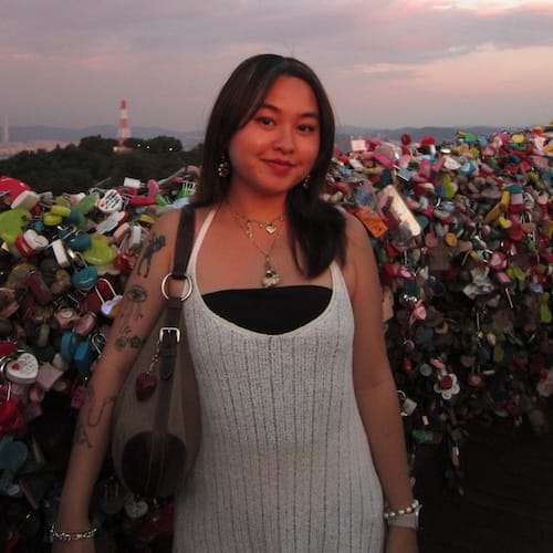 Thavon Her has long dark hair and is wearing a white halter dress, large pendant necklace and is holding a bag under her right arm. She is standing on a bridge covered with colorful locks during sunset.