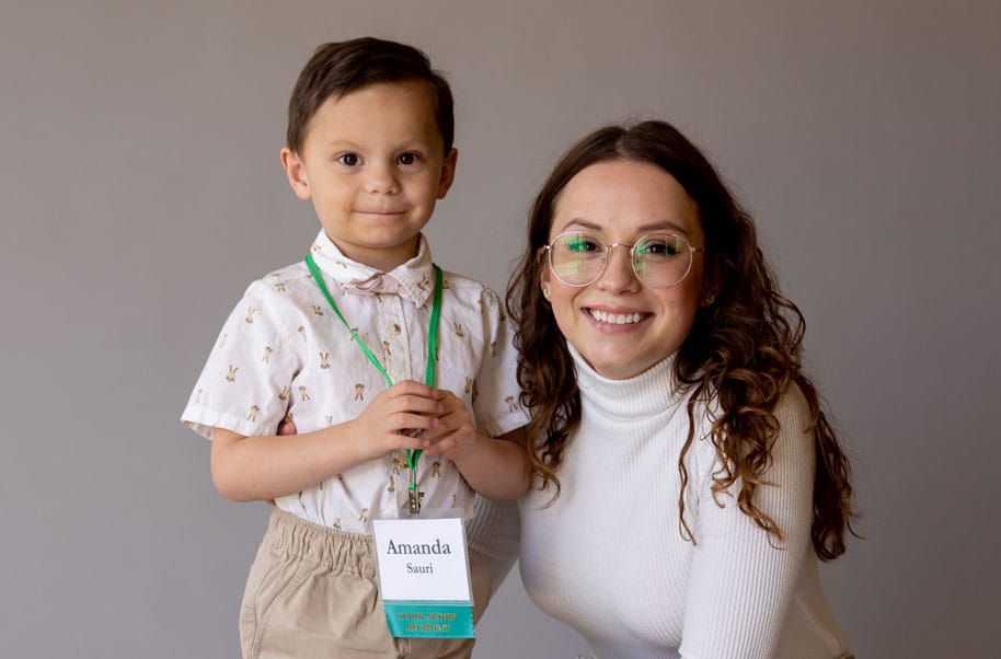 Amanda Sauri kneels next to her young son against a light brown backdrop