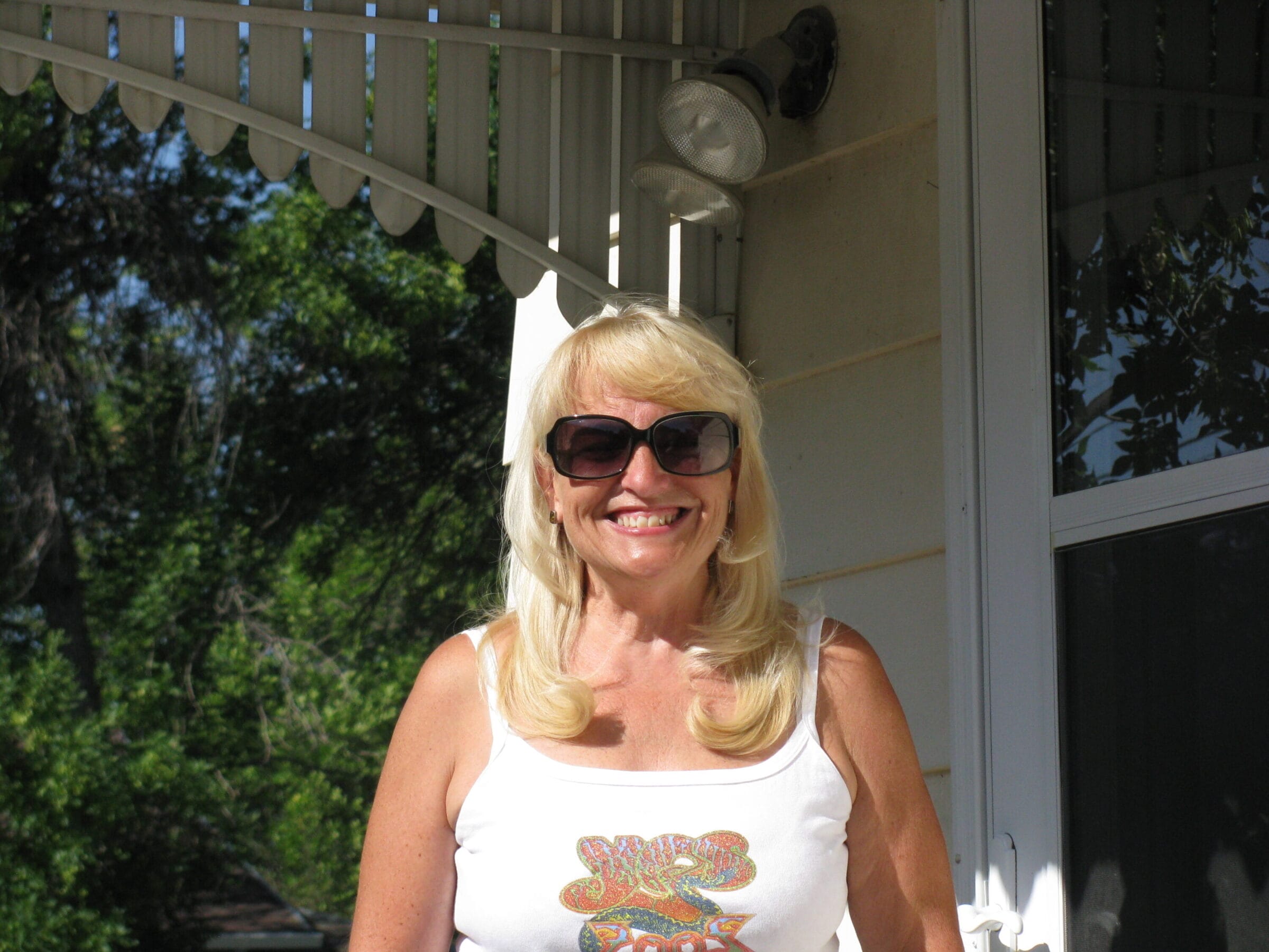 Jeannette Eichholz stands in a tank top and sunglasses on her front porch on a sunny day