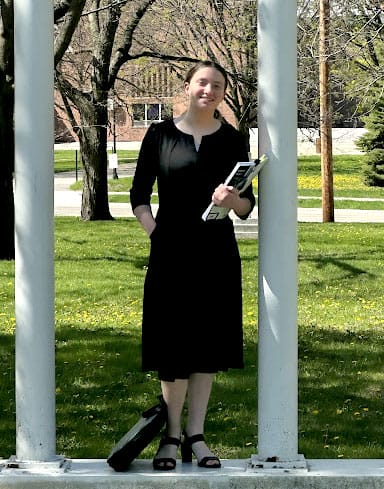 Alina Savagian-DeMoss stands outside between two white pillars, holding a book and smiling.