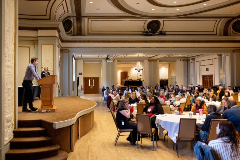 A speaker stands at a podium addressing a large audience seated at round tables in Memorial Union’s Great Hall.