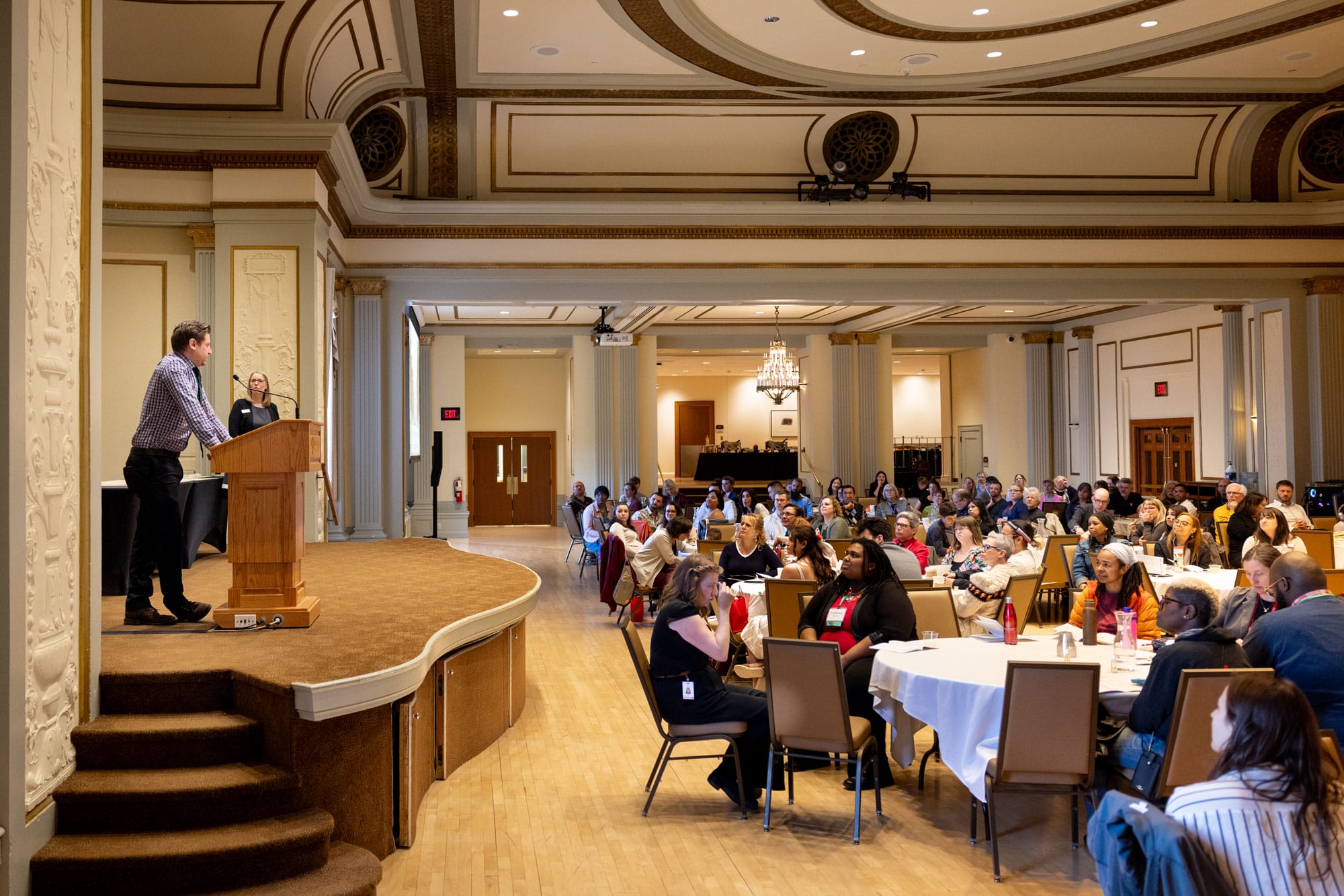 A speaker stands at a podium addressing a large audience seated at round tables in Memorial Union’s Great Hall.