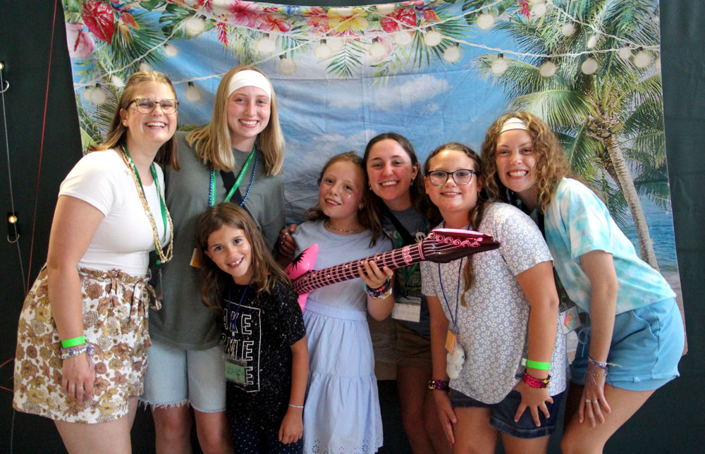 Three young women and four young girls standing and smiling in front of a tropical-themed backdrop. One is holding a guitar.