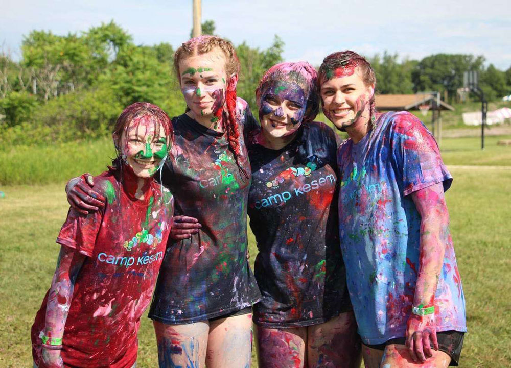 Four young women at a summer camp, standing in a field, splattered with paint, with their arms around each other and smiling.