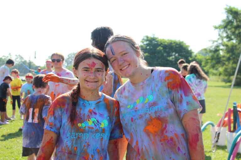 Two young women splattered with paint, standing in a field and smiling at the camera.