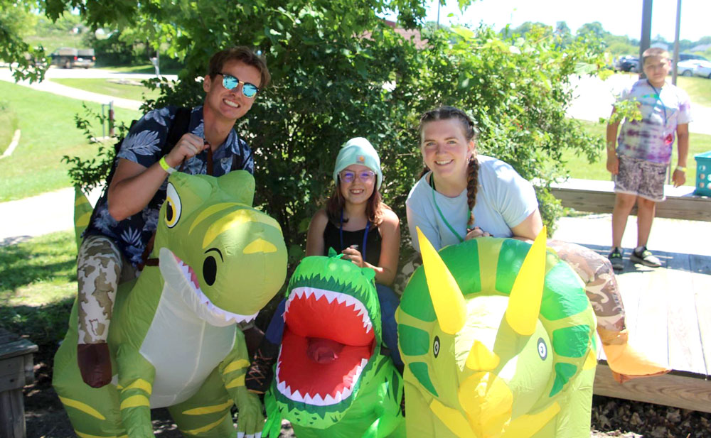 Three young people wearing green and yellow dinosaur costumes that go from the waist down, standing in front of a tree and smiling.