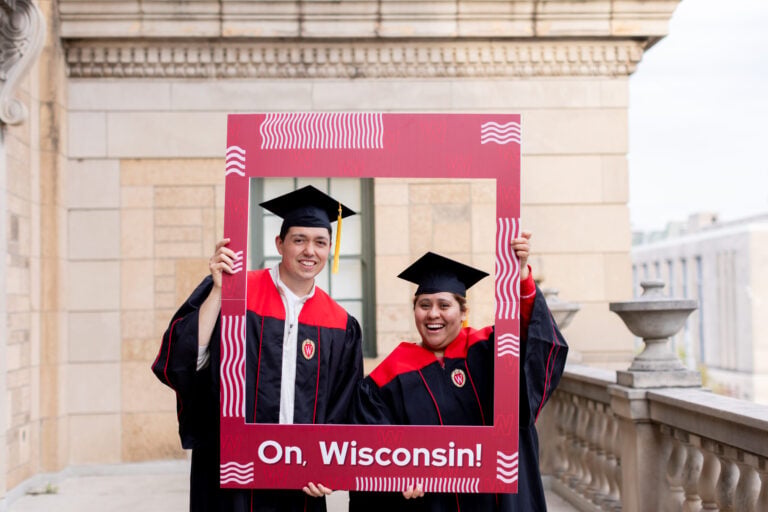 Adult students in cap and gown holding a cardboard photo frame that says, “I’m a Badger.”