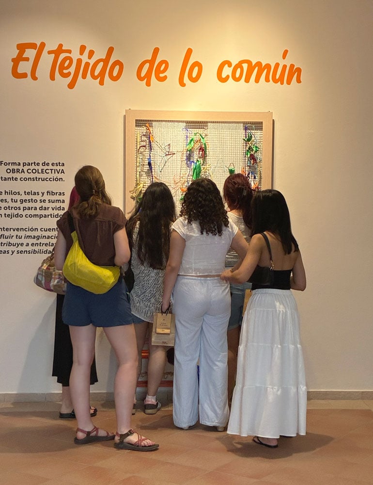 A small group of students facing away from the camera look closely at a wall-mounted artwork.