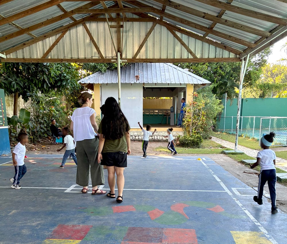 Children play in a covered outdoor space. Two UW–Madison students stand in the middle of the space, faces turned away.