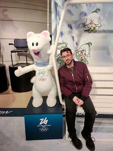 Man sitting next to Olympics mascot, a white stuffed animal mascot called “Tina.”