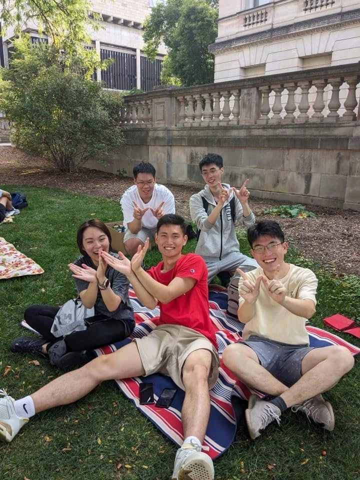 Five young adults sit on a blanket on grass, smiling and making hand gestures.