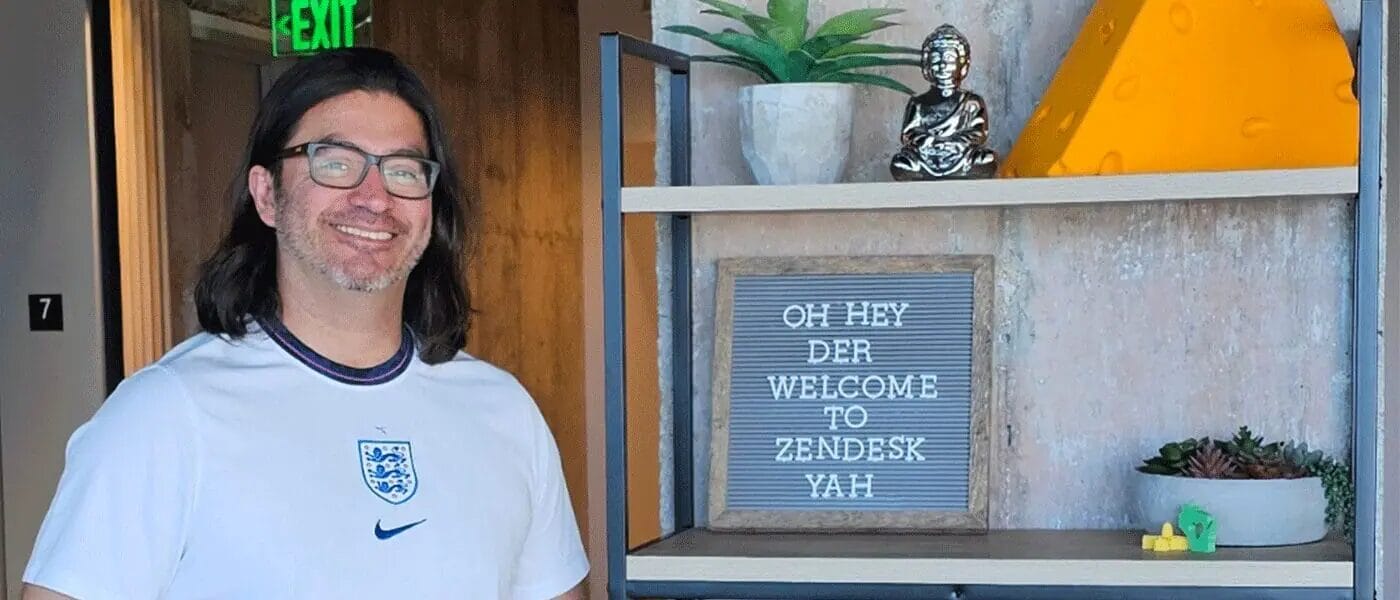 Man in England soccer jersey smiles next to a shelf with a welcome sign and decorative items.