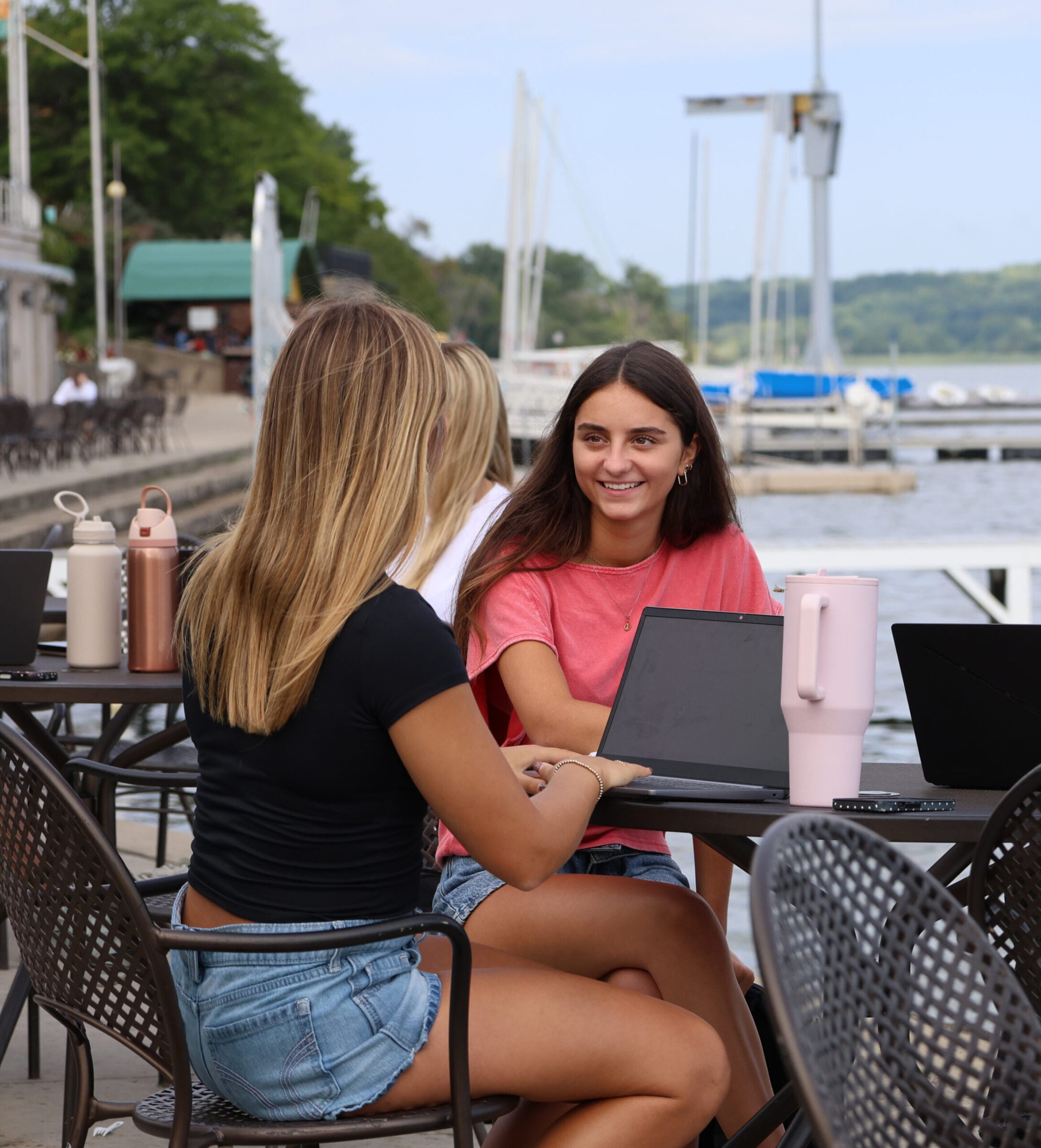 Two young women with laptops at a table by the water.